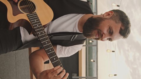 Handheld vertical shot of happy bearded young man playing acoustic guitar on rooftop terrace on nice summer evening