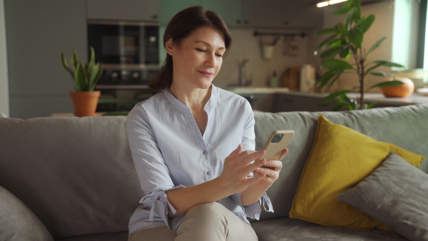 Happy middle aged mature woman using smartphone texting message sit on couch, lady hold phone typing sms enjoying communication in mobile app at home, older people and modern tech concept. Mothers Day Royalty-Free Stock Footage #1076652899