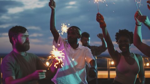 Handheld slowmo shot of multi-ethnic group of happy young people with sparklers partying together on rooftop terrace on summer evening