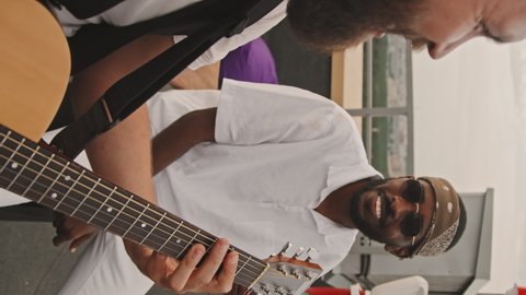 Vertical shot of cheerful bearded Caucasian young man playing acoustic guitar and smiling while his African-American male friend drinking beer and enjoying music at rooftop party