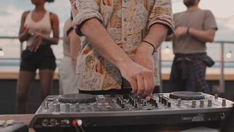 Handheld close up shot of unrecognizable DJ playing set at rooftop party while young people dancing in background