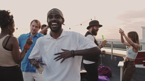 Handheld shot of happy young African-American man with cocktail dancing and looking at camera at rooftop party on summer eveningの動画素材