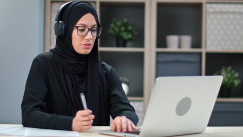 Smiling modern Muslim freelancer business woman talking online video call laptop working remotely at home office. Happy Asian lady in black hijab studying internet distance education on computer app