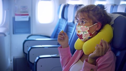 a bright modern young European woman is on an airplane waiting for a take off and inserting earplugs in her ears. a woman in a pink shirt with a neck pillow wearing an unusual fashionable protective
