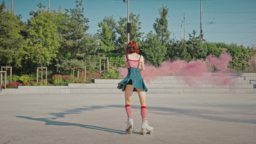 Outdoor dances. Young beautiful pin up lady roller skating with pink smoke bomb outdoors, dancing gracefully in urban park, tracking shot, slow motion