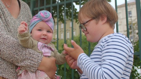 Down syndrome boy touches and examines baby toddler feet. Acquaintance with new, knowledge world girl in mother arms rejoices at the attention. CZ, Prague, 20.7.2021.
