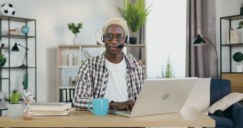 Portrait of lovely satisfied smiling young black-skinned gay with blond hair in headset which sitting in front of camera in home office during work on laptop,LGBT-community concept