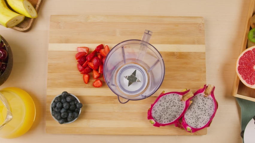 Woman hands putting ripe strawberries and blueberries into blender close-up, cooking smoothie, healthy sweet cocktail without sugar. Fresh fruits, halves of pitahaya on kitchen table. 