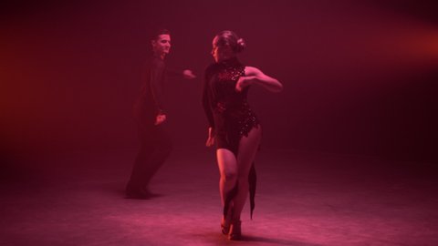 Full length ballroom dancers performing cha cha cha indoors. Young dance couple taking part in ballroom competition inside. Attractive man and woman dancing latina in red light background.