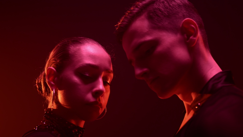 Closeup dance couple looking each other in red light background. Side view young dancers holding hands indoors. Ballroom partners dancing inside. 