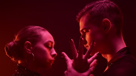 Closeup dance couple looking each other in red light background. Side view young dancers holding hands indoors. Ballroom partners dancing inside. 