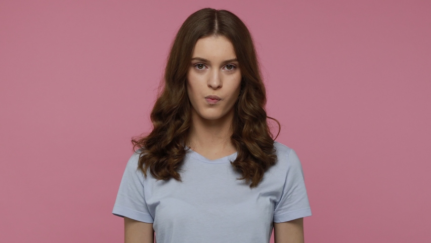 Portrait of stunning teenager girl with wavy hair in blue casual T-shirt looking camera, covering her eye bitcoin, future currency. Indoor studio shot isolated over pink background.