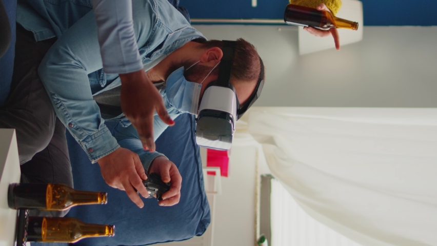 Vertical video: Multiethnic friends enjoying vr tehnology playing video games in living room wearing mask to prevent infection with coronavirus keeping social distancing. Diverse people having fun at