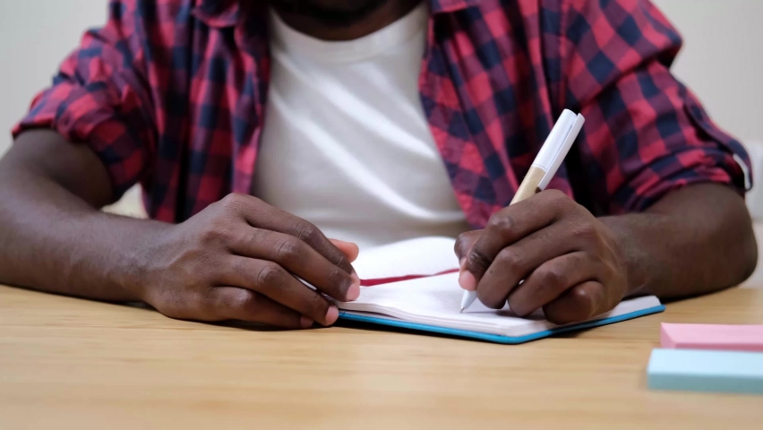 Close-up shot of biracial black man writing down notes in paper notebook while sitting at the table. Young entrepreneur, businessman assigning daily tasks in a personal organizer.