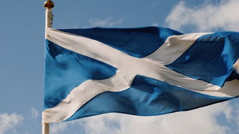 Slow motion view of the flag of Scotland, also known as St Andrews Cross, flying against a blue sky background with white volumetric clouds