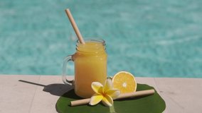 Video footage of glass mason jar with orange juice, bamboo straw, half of fresh orange, yellow frangipani flower and bubbling blue swimming pool on background.