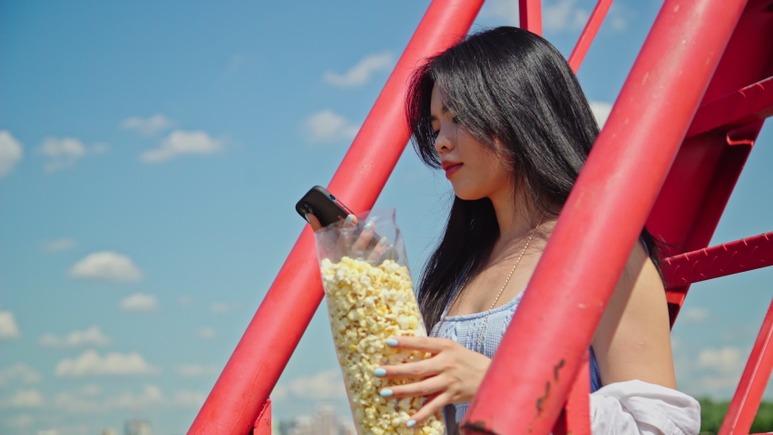 Beautiful happy young asian woman talking on the phone with popcorn while sitting on the red stairs in the park on a sunny day.Girl in casual outfit outside. Attractive lady with dark hair 4K