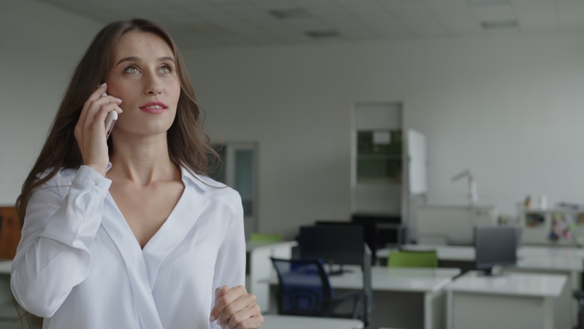Beautiful young lady stands and speaks on her black phone, white shirt, office