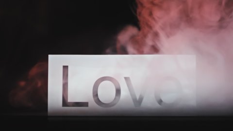 Close up slowmo shot of sign with word Love isolated on black background. Wisps of red smoke falling on it from above and dissipating