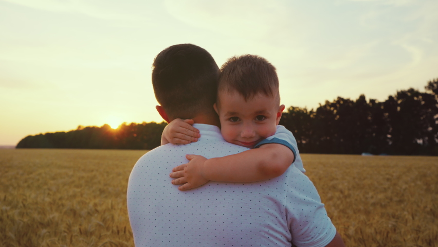 Father holding little son in arms and walking towards setting sun in wheat field, boy looking at camera and hugging his dad. Family spending time together. Concept of love Royalty-Free Stock Footage #1076715515