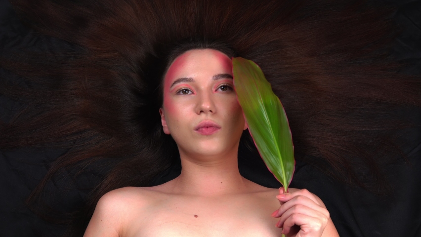 Beautiful young model posing for a photographer. A fashionable model with bright makeup holds a green leaf in her hands. Black fabric background. 4k footage