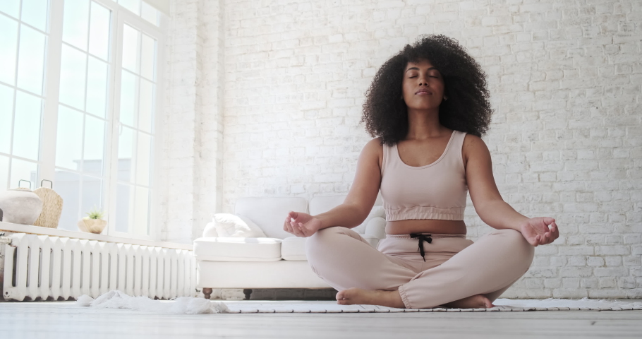 A black woman sits in a lotus position. Place hands on lap, fingers folded, eyes closed, and meditate. Doing yoga at home. Anxiety reduction concept, no stress, healthy habit, mindfulness lifestyle. Royalty-Free Stock Footage #1076719124