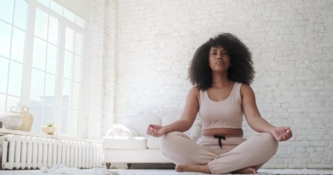 A black woman sits in a lotus position. Place hands on lap, fingers folded, eyes closed, and meditate. Doing yoga at home. Anxiety reduction concept, no stress, healthy habit, mindfulness lifestyle.