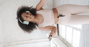 A happy mixed-race woman wearing headphones enjoys moving. Relaxing in the bedroom while listening to music, a cheerful young lady with earbuds dances enjoys a carefree weekend at home. Vertical video
