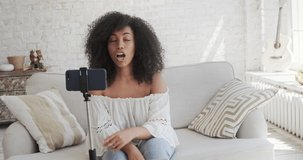 Young mixed-race blogger streaming online in a social network and communicates with her subscribers, recording video vlog podcast on smart phone sitting at home, chatting on a video call