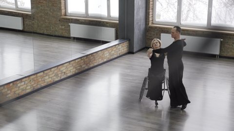 Top view of positive middle aged dancers, attractive blonde woman with differing abilities on wheelchair and imposing man dancing hand in hand. Graceful pair whirling holding hands in dance hall