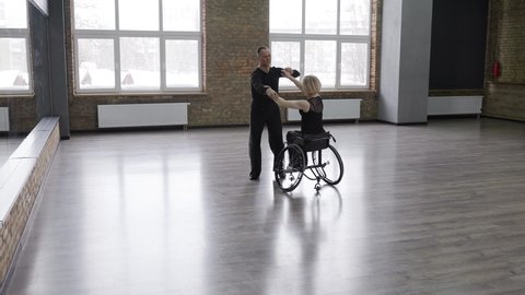 Attractive couple of professional ball dancers during training, man whirling handicapped woman sitting on wheelchair. Handicapped female and her partner preparing for competitions in ballroom