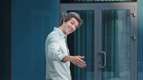 Portrait of smiling successful friendly young man in formal attire standing by door, hr recruiter stretching hand to camera glad to offer you job in company, greeting spectator as new workforce member