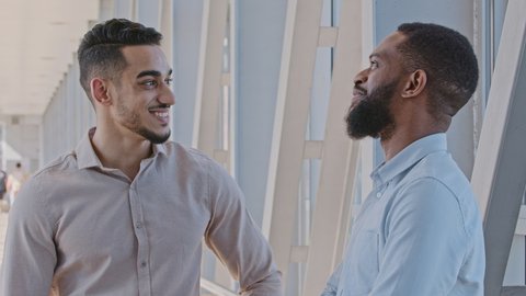 Two multiracial colleagues business men african american guy and hispanic man standing in hallway in office talking communication conversation informal handshake male hug support congratulation