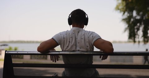 Rear view man puts on big headphones sitting on bench, slow motion