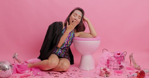 Sleepy but happy Asian woman yawns and has fun after party leans on toilet bowl wears fashionable dress black jacket poses in lavatory room recalls music from disco makes rock n roll gesture