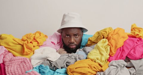 Displeased dark skinned man covered with laundry wears panama has dissatisfied expression cluttered with clothes being fully buried and unrecognizable. White background. Domestic work concept