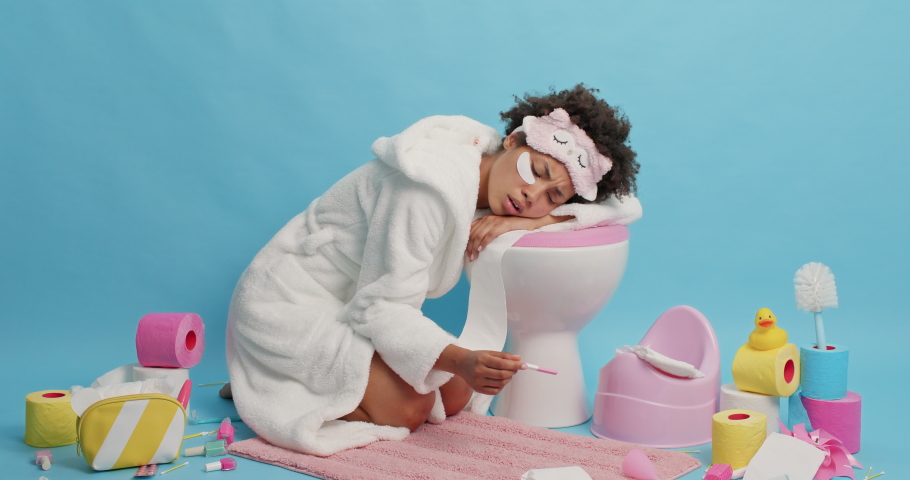Unhappy curly haired woman leans on toliet bowl dissatisfied with pregnancy result wears white bathrobe and sleepmask has stressed expression isolayed over blue background. Poor contraception