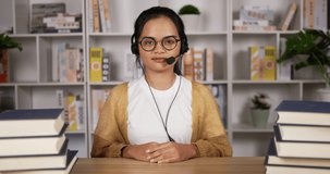 Smiling asian female student in headphones and glasses sit at desk at home. Happy young woman in earphones talking at camera. Learn or work from home. Girl looking at camera.