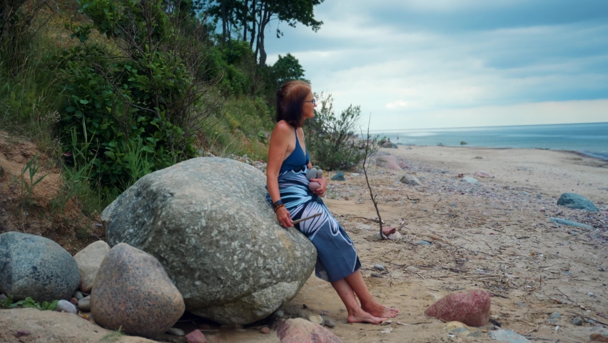 Old Woman Sitting On A Huge Rock By The Beach In Karkle, Lithuania - wide shot
