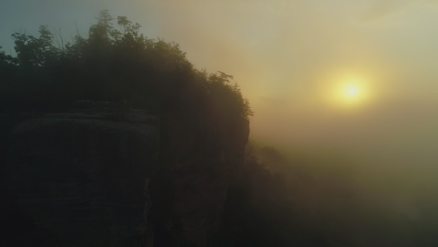 Aerial orbiting two silhouettes on cliff overlook during foggy golden sunrise, 4K