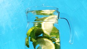 Lemonade in a transparent jug with rotating sliced lemons, limes and mint leaves close-up on a light blue background. 4k slow motion video.