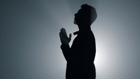 Silhouette of young man whispering prayer in dark space. Religious male person praying in spotlights background. Believer human turning to god with folded hands indoors. 
