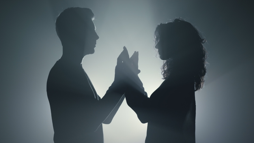 Silhouette of romantic couple standing opposite each other in spotlight background. Closeup sensual man and woman holding hands in dark space. Young lovers crossing arms indoors. 