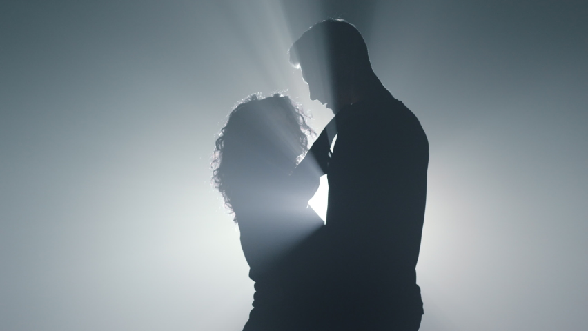 Silhouette of sensual lovers hugging in darkness. Romantic couple touching each other in spotlights background. Sexy man and woman dancing slowly indoors. 