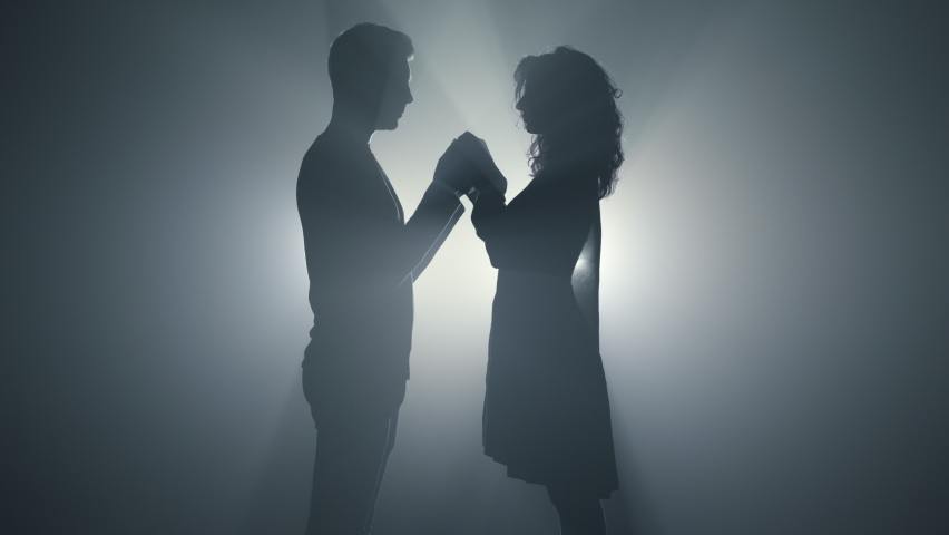 Silhouette of loving couple holding hands in floodlight background. Beautiful guy and girl standing in dark space. Sensual lovers spending time together indoors. 