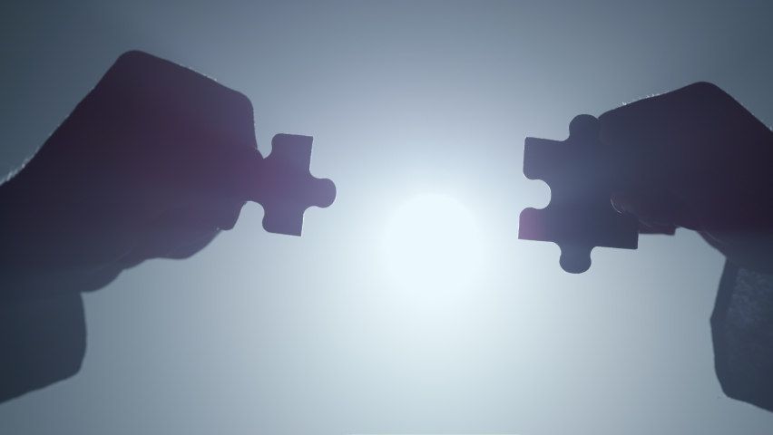 Silhouette of man and woman arms holding pieces of jigsaw in floodlight backdrop. Closeup two hands connecting parts of puzzle in darkness. Unknown couple matching elements into whole indoors. Royalty-Free Stock Footage #1076743328