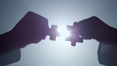 Silhouette of man and woman arms holding pieces of jigsaw in floodlight backdrop. Closeup two hands connecting parts of puzzle in darkness. Unknown couple matching elements into whole indoors.