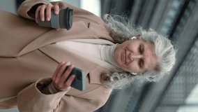Vertical video. Stylish mature woman enjoying hot coffee outdoors and using wireless earphones and smartphone. Business lady with modern gadgets on street.