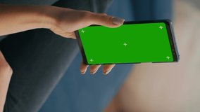 Vertical video: Freelancer holding in vertical mode smartphone with mock up green screen chroma key display while lying on sofa in living room. Woman using isolated touchscreen device for social
