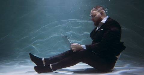 Cinematic under water side view, young happy workaholic businessman in black suit using laptop to work slow motion.